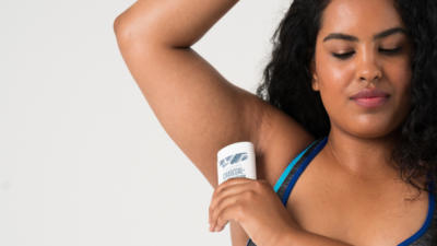 Woman with brown curly hair wearing sports bra looking down applying Schmidt's Charcoal + Magnesium natural deodorant to her underarm in front of a grey background